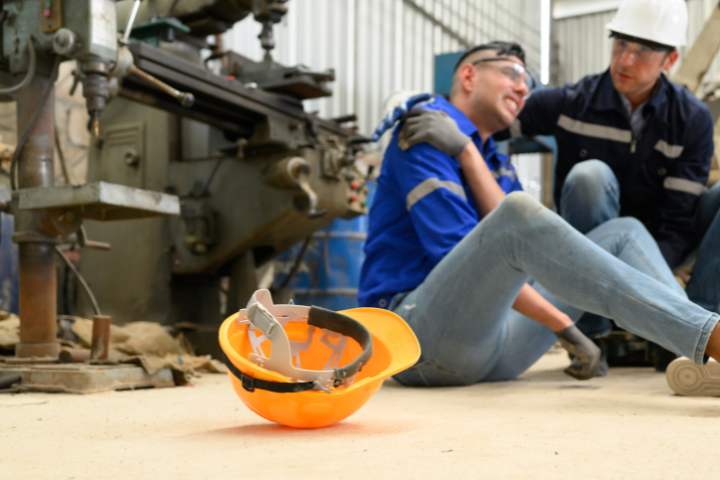 Trabalhador acidentado sendo amparado por colega em ambiente industrial, capacete de proteção no chão, paleta azul e cinza, fotografia realista dramática
